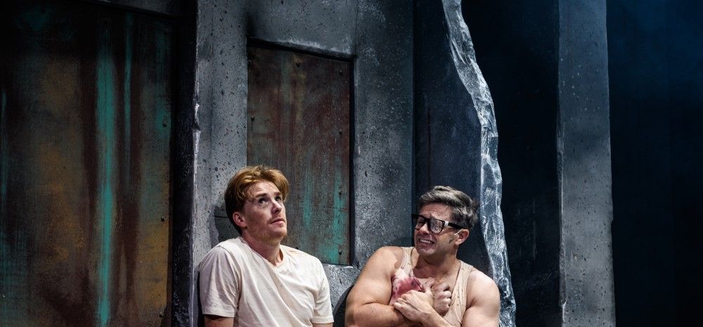 Two male actors sit on a bench against a dark, industrial set. They wear simple T-shirts and shorts, barefoot, with a distressed and tense expression, suggesting a prison or interrogation setting