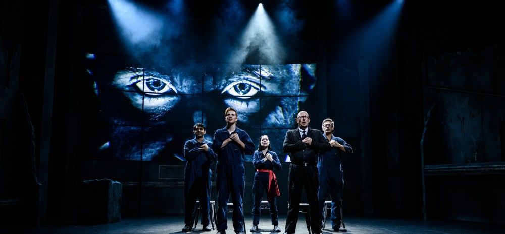 Five actors stand in a row on stage in dark jumpsuits, hands clasped at their chests. Behind them, a large screen displays a close-up of intense, watchful eyes, lit with dramatic blue spotlights.