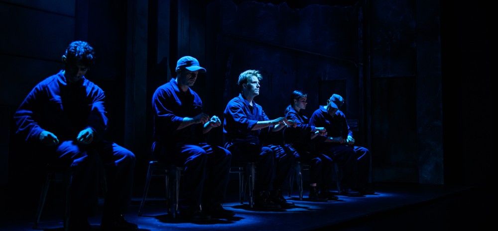 Five actors sit in a line, illuminated by low blue stage lighting, miming mechanical tasks with their hands as if operating invisible controls. They are dressed in dark jumpsuits.