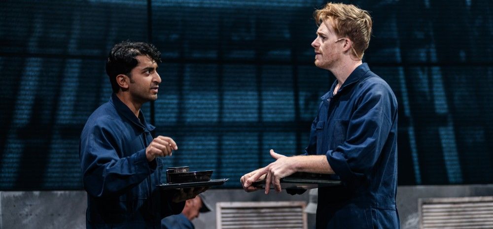 Two actors in navy blue jumpsuits stand in conversation holding metal trays, as if in a canteen. The background features an industrial set design with other performers sitting and eating.
