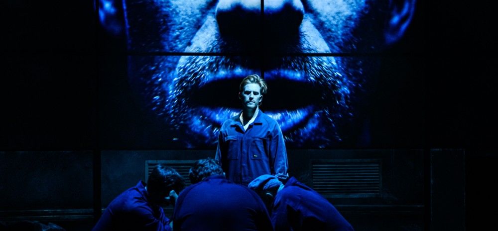 A central actor in a jumpsuit stands under harsh blue light, surrounded by others hunched over a table. Behind them, a giant projected face stares out, creating an ominous atmosphere.